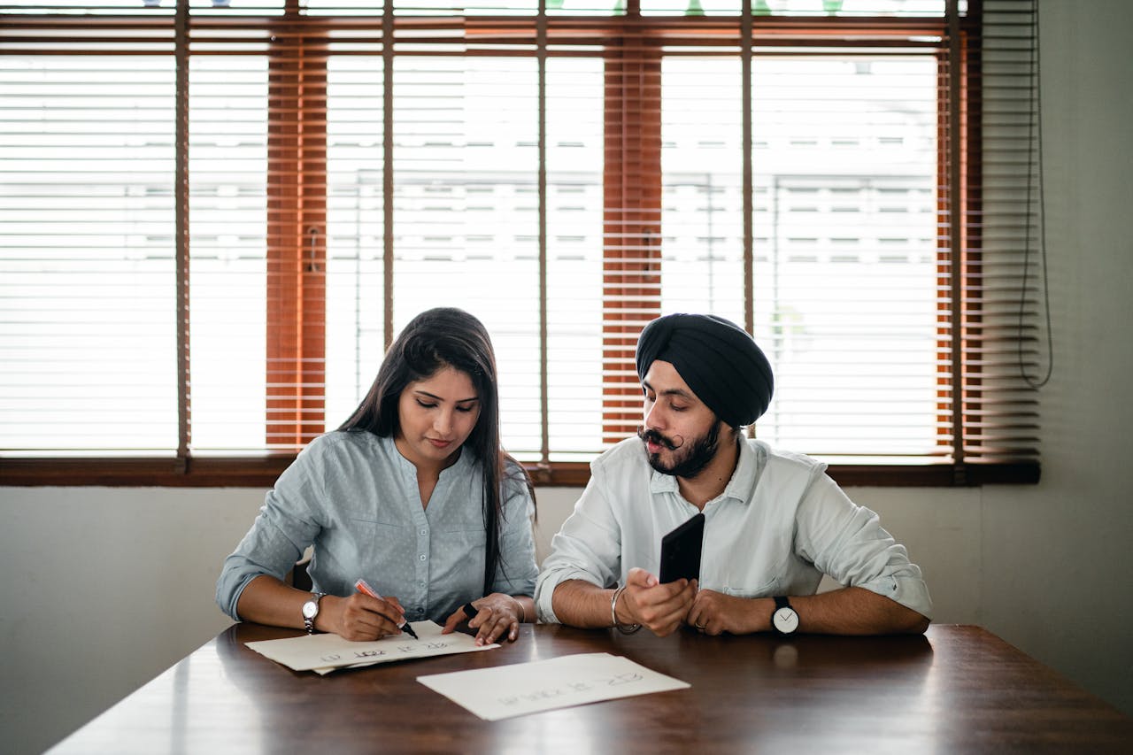 services-06 Indian woman with papers and pen writing on paper poster with ethnic Indian man in turban sitting nearby at wooden table
