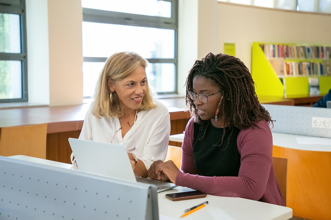 Crafting Captivating Headlines: Your awesome post title goes here Positive adult female teacher explaining task to young black student with Afro braids doing assignment on laptop in modern classroom